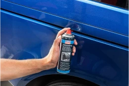 Hand sprüht Lackpflege-Spray auf blaue Autotür, Dose nah am Fahrzeugblech gehalten.