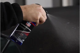 Hand sprüht Autopflege-Spray auf eine schwarze Lackfläche, feiner Nebel verteilt sich gleichmäßig.